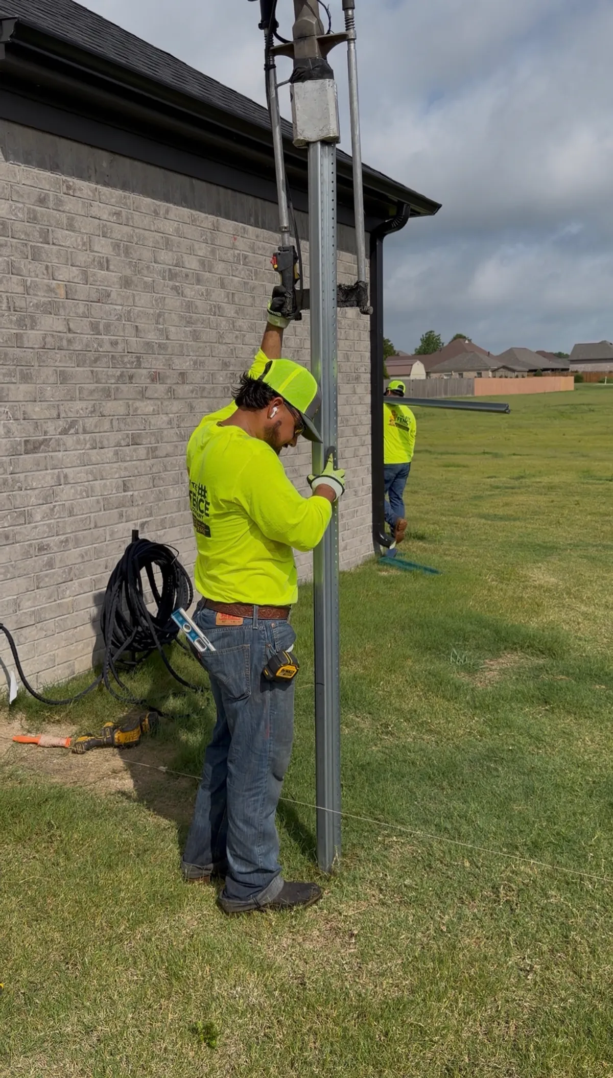 The Fence Company crew driving a galvanized steel fence post into undisturbed soil beside a brick home